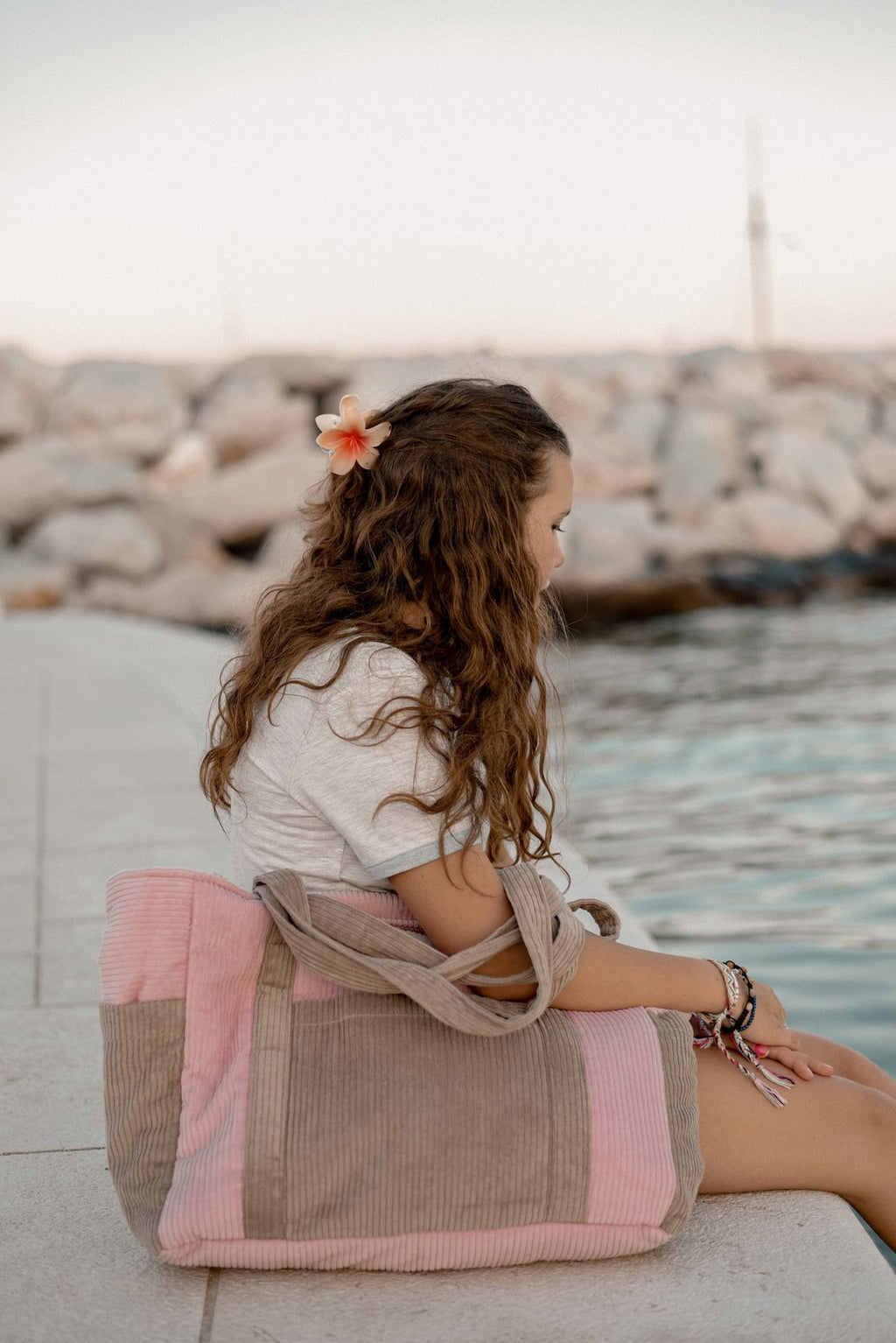 2. Woman sitting by the water with pink and coffee-colored weekender bag beside her, wearing a flower in her hair