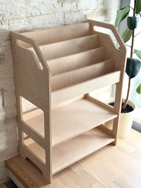 1. Montessori wooden book display with storage shelf in natural birch plywood next to a potted plant in a bright room