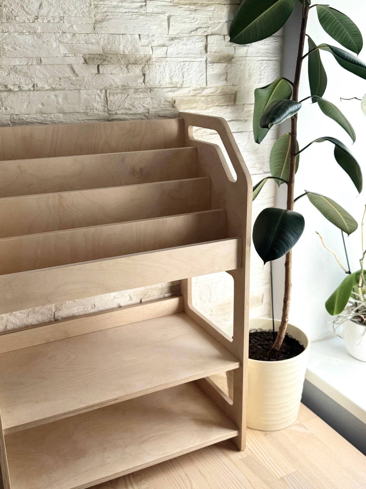 1. Montessori wooden book display with storage shelf in natural birch plywood next to a potted plant in a bright room