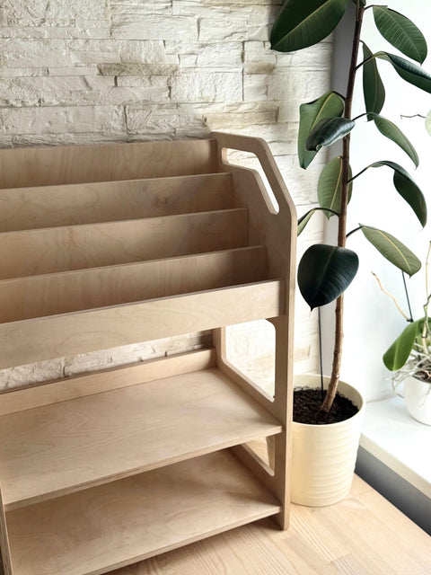 1. Montessori wooden book display with storage shelf in natural birch plywood next to a potted plant in a bright room