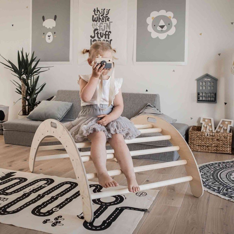 1. Young girl sitting on wooden Montessori climbing arch in a cozy playroom, holding a toy camera, with playful wall art in the background