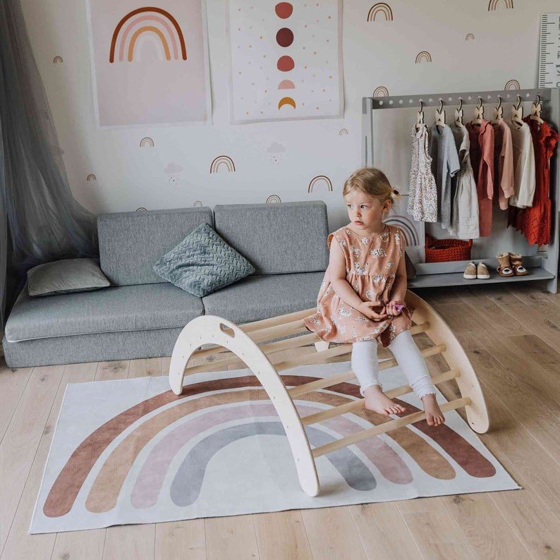 2. Girl in a peach dress sitting on a wooden Montessori climbing arch in a colorful playroom with rainbow-themed decor