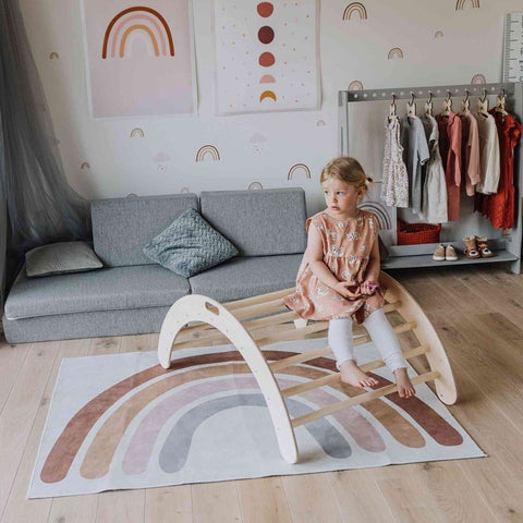 2. Girl in a peach dress sitting on a wooden Montessori climbing arch in a colorful playroom with rainbow-themed decor