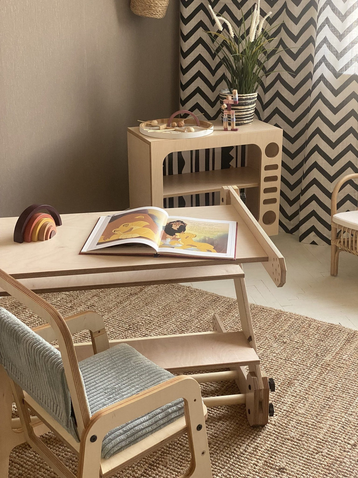 9. Montessori table setup with a wooden chair and open book, in a playroom with a chevron curtain and decorative shelf.