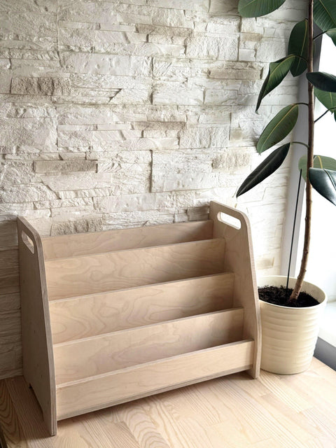 1. Montessori wooden book display with four tiers in a modern room setting next to a potted plant, showcasing its natural birch plywood finish and minimalist design.