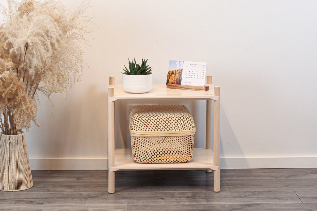 1. Montessori-inspired wooden shelving unit with two shelves, featuring a potted plant and a calendar on top, next to a vase with dried flowers, set against a white wall and wooden floor.