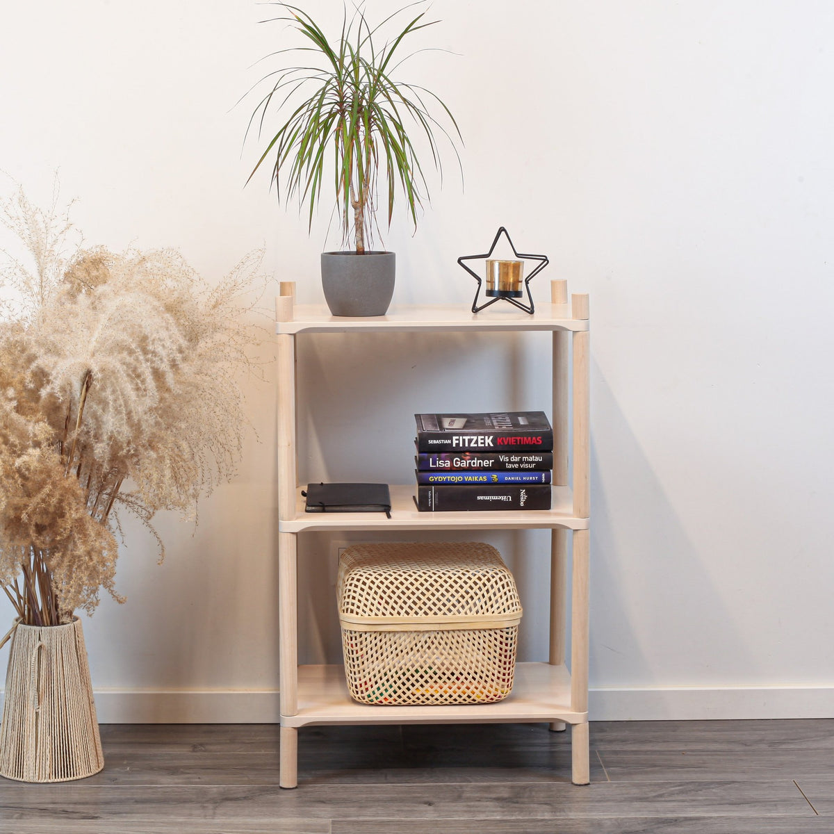 1. Montessori-inspired wooden shelving unit with three shelves, featuring a potted plant, books, and a decorative star on top, set against a white wall with pampas grass on the side