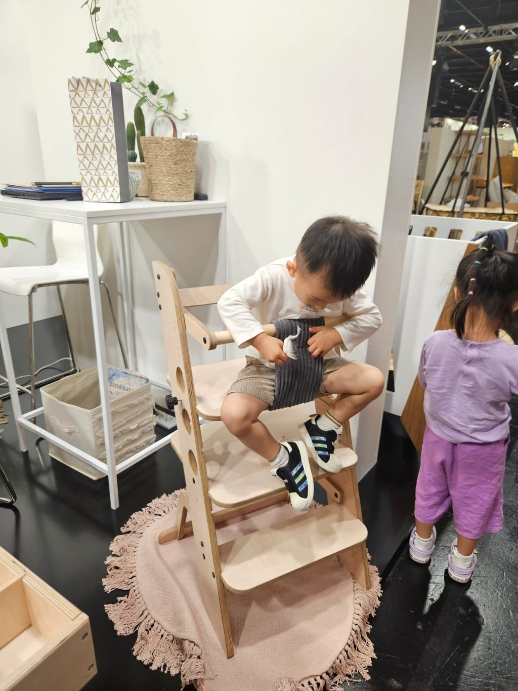 1. Child sitting on wooden Montessori learning tower in a playroom, with another child nearby