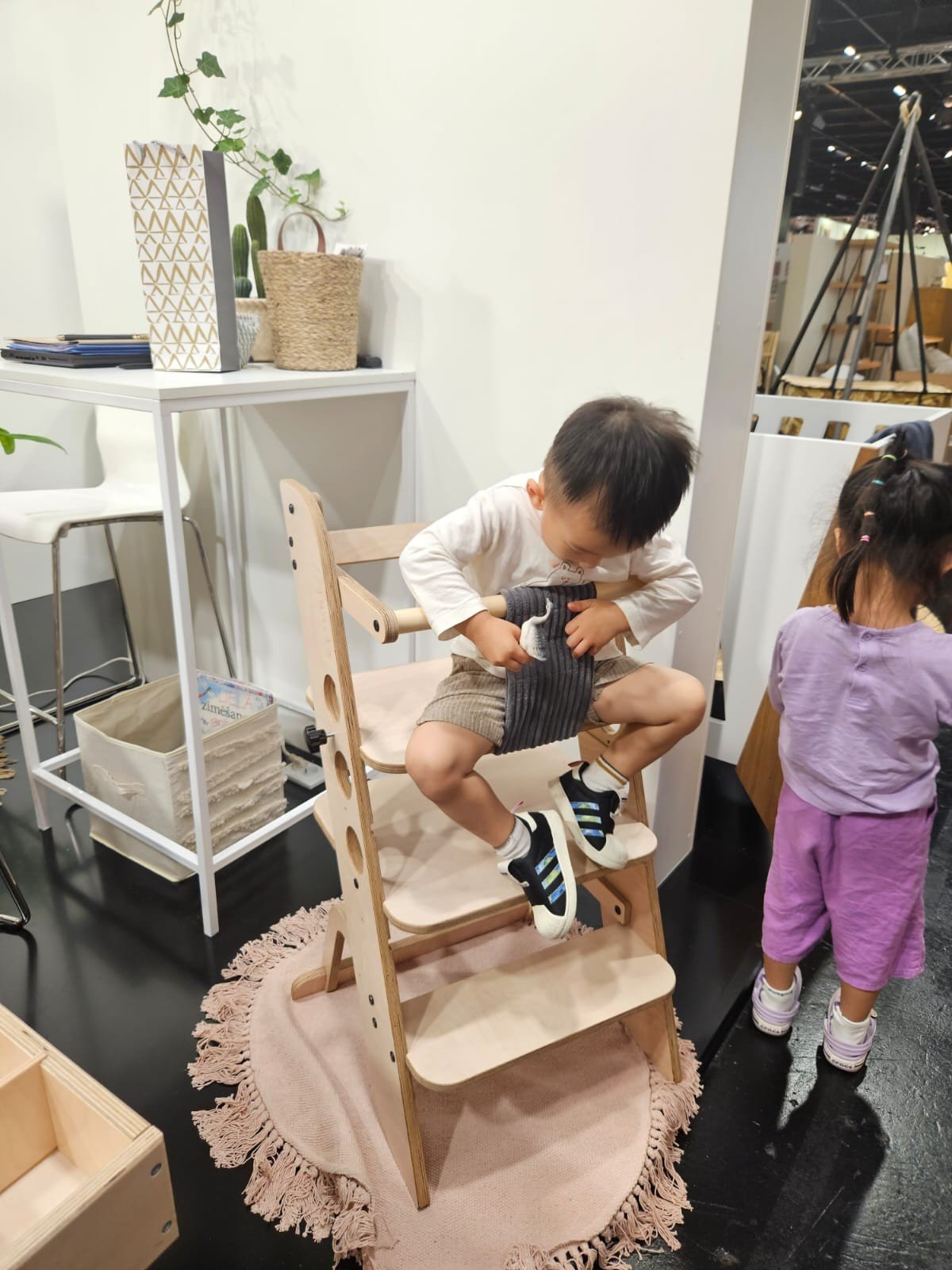 1. Child sitting on wooden Montessori learning tower in a playroom, with another child nearby