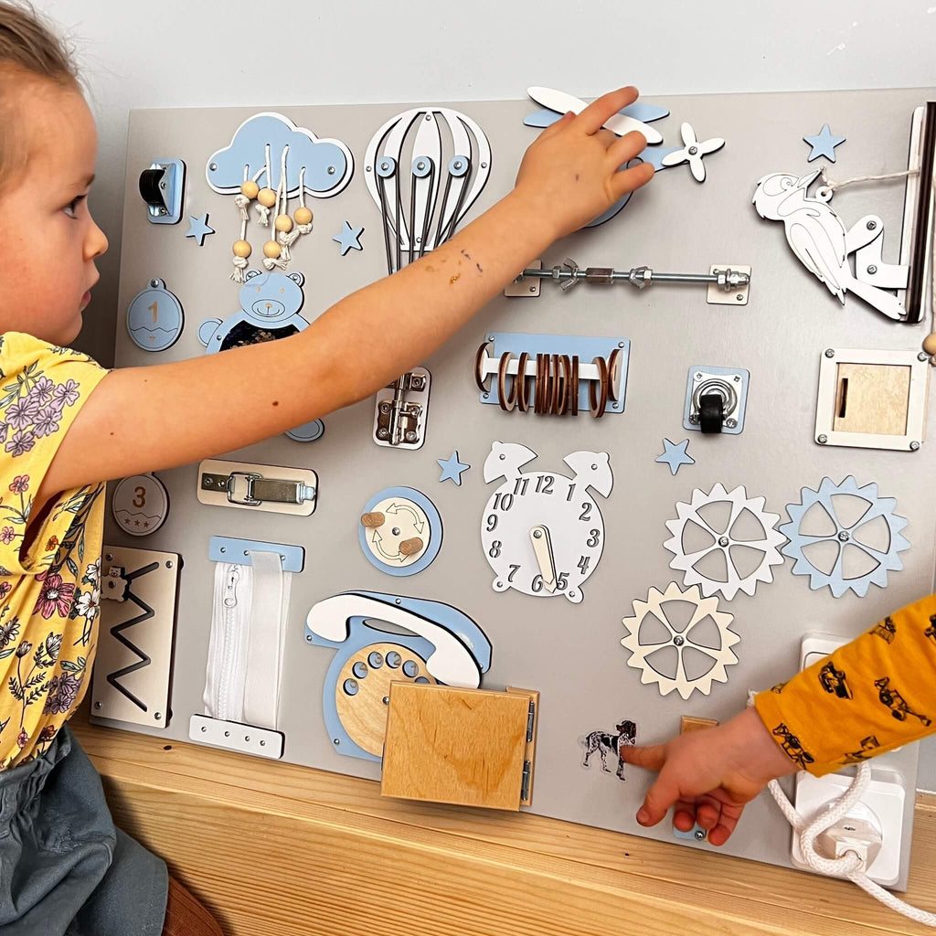1. Two children playing with a Montessori busy board featuring blue and white elements, designed for interactive skill building.