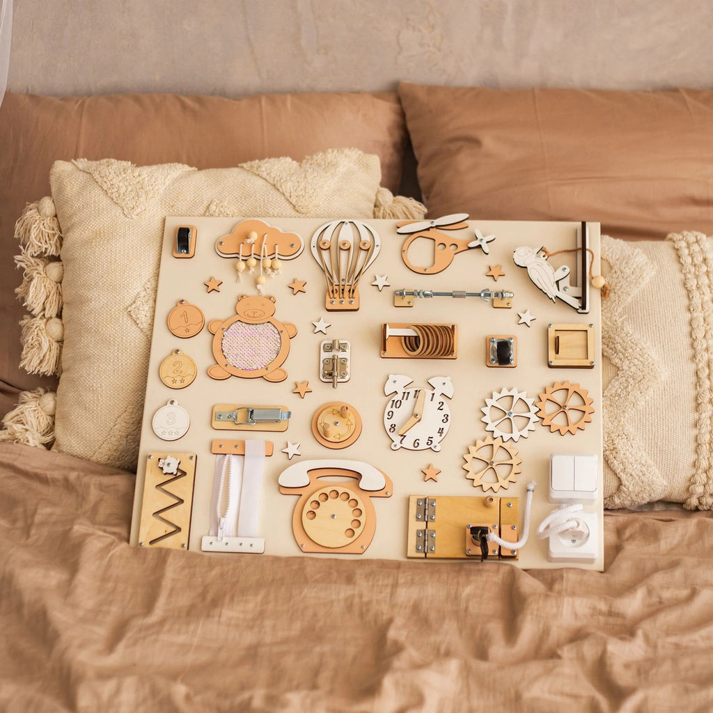 1. Montessori busy board with beige and orange elements on a bed with textured pillows, showcasing interactive features for children's play.