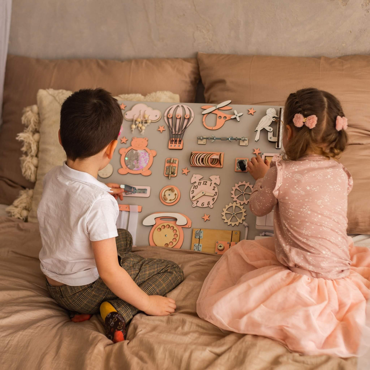 1. Two children playing with a Montessori busy board featuring orange and beige elements on a bed, designed for interactive learning.