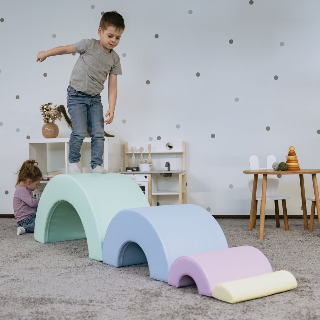 1. Boy balancing on pastel rainbow soft play set in playroom with girl playing in background