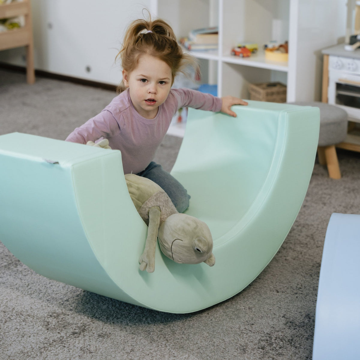 8. Girl sitting on pastel rainbow soft play piece with stuffed toy in playroom
