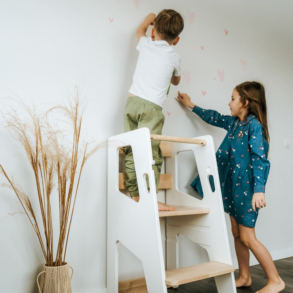 1. Two children using white Montessori Tower Safety+ to reach wall decorations, highlighting its safety and collaborative use