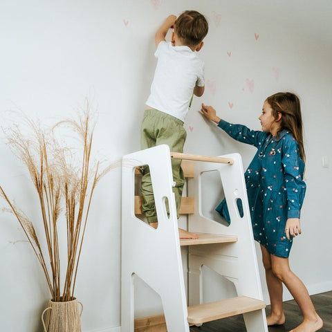 1. Two children using white Montessori Tower Safety+ to reach wall decorations, highlighting its safety and collaborative use