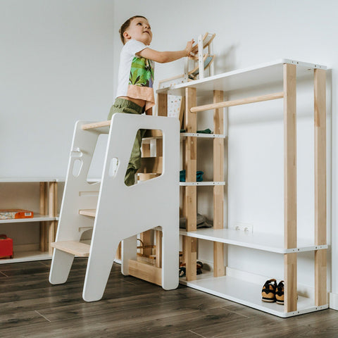 1. Child climbing white Montessori Tower Safety+ next to a wooden shelf, emphasizing its integration into home activities