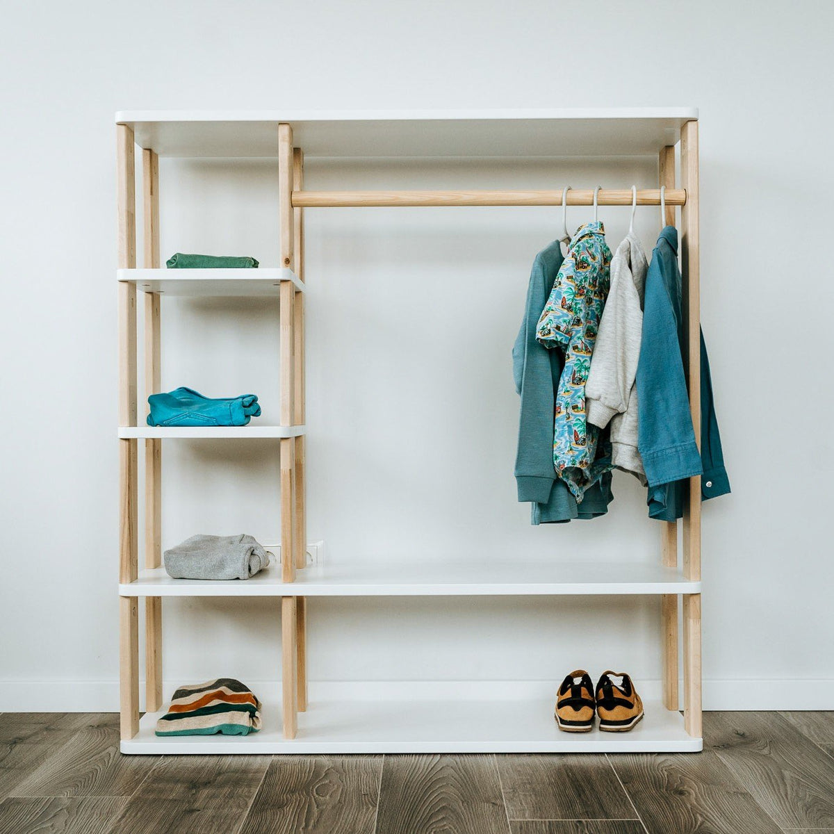 1. Montessori wardrobe with white shelves and natural wood clothes rail, displaying children's clothing and shoes, set against a white wall on wooden flooring