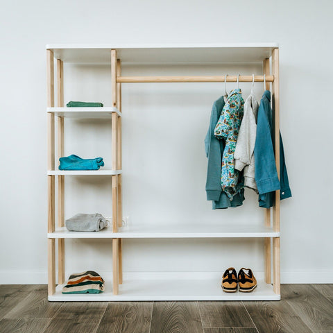 1. Montessori wardrobe with white shelves and natural wood clothes rail, displaying children's clothing and shoes, set against a white wall on wooden flooring