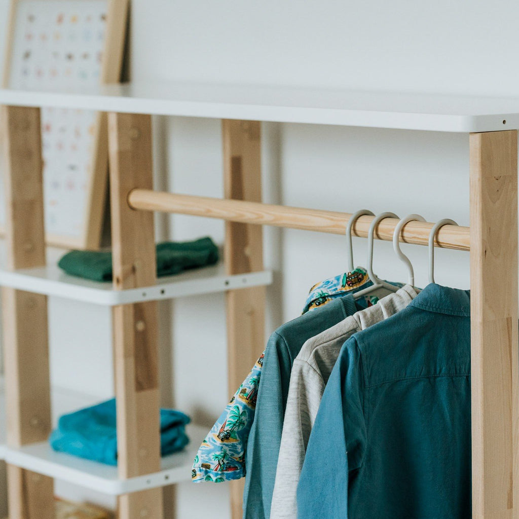 3. Close-up of Montessori wardrobe's natural wood clothes rail with children's shirts hanging, highlighting shelf detail and clothing texture