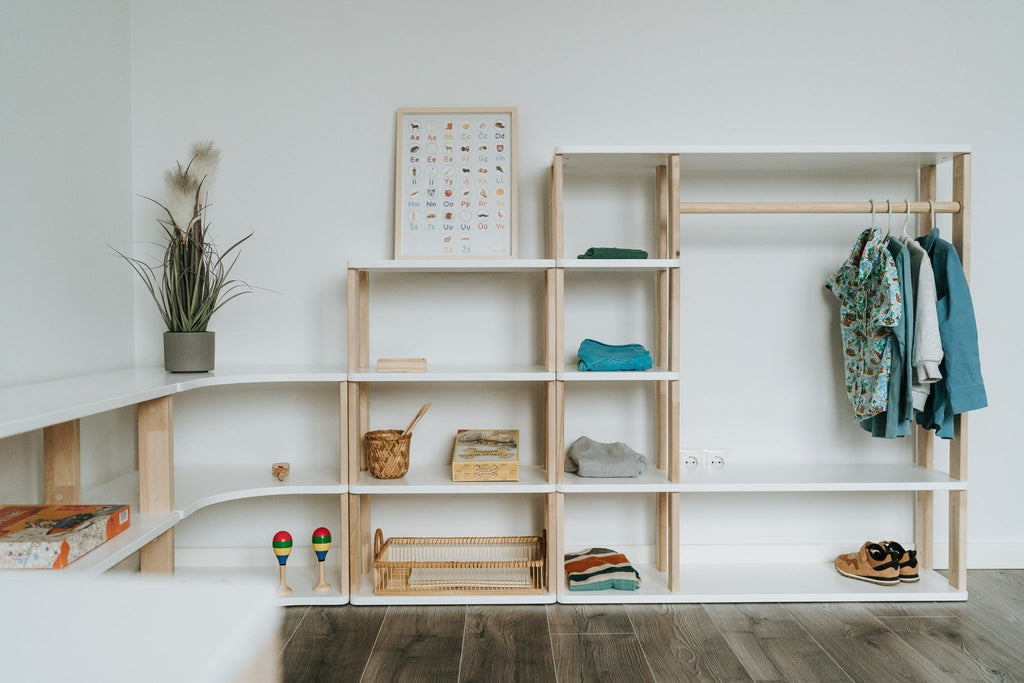 2. Montessori wardrobe and shelving unit in white and natural wood, featuring children's clothing and toys, set against a white wall