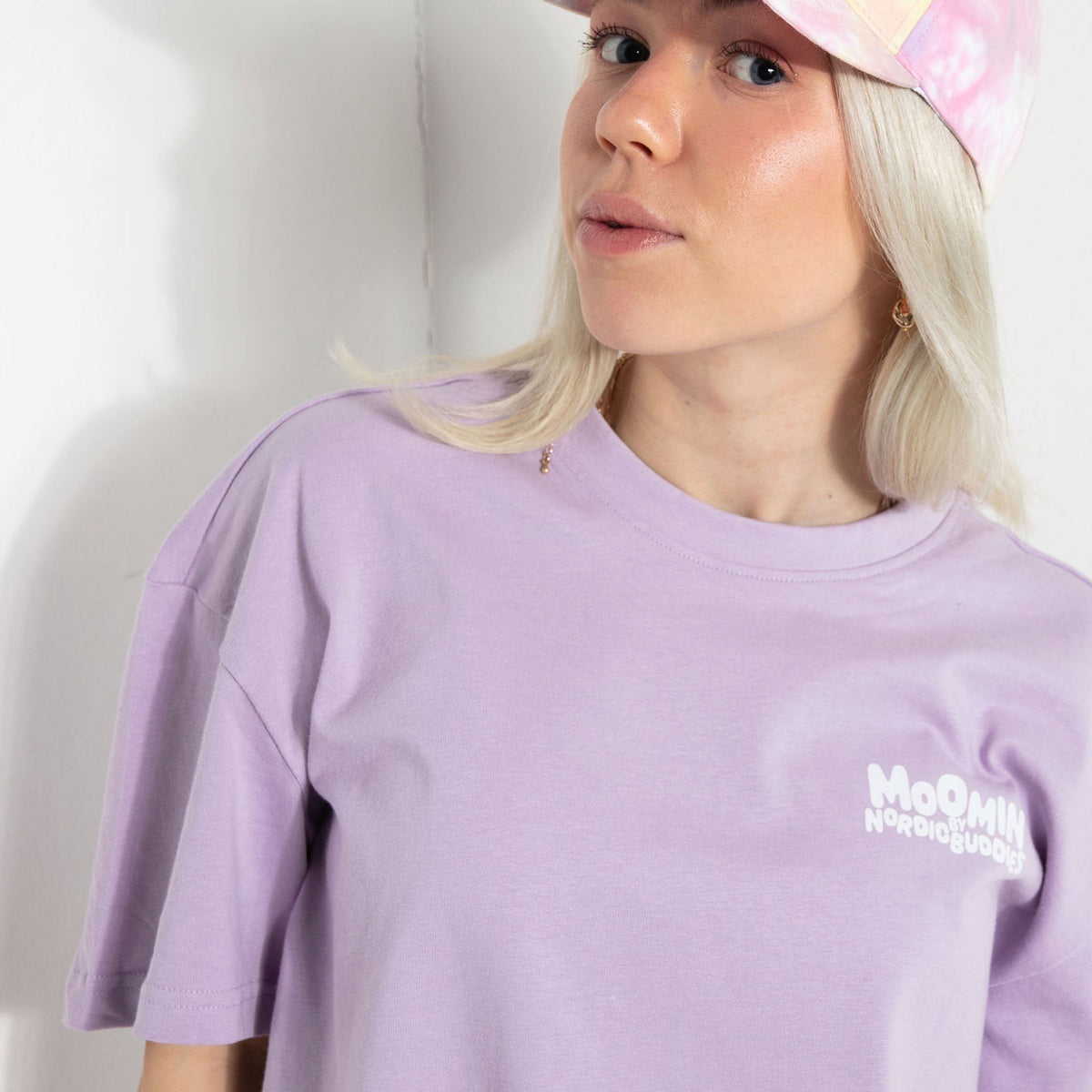 1. Female model wearing lilac Moomin unisex t-shirt with white logo, paired with pink cap, in casual setting
