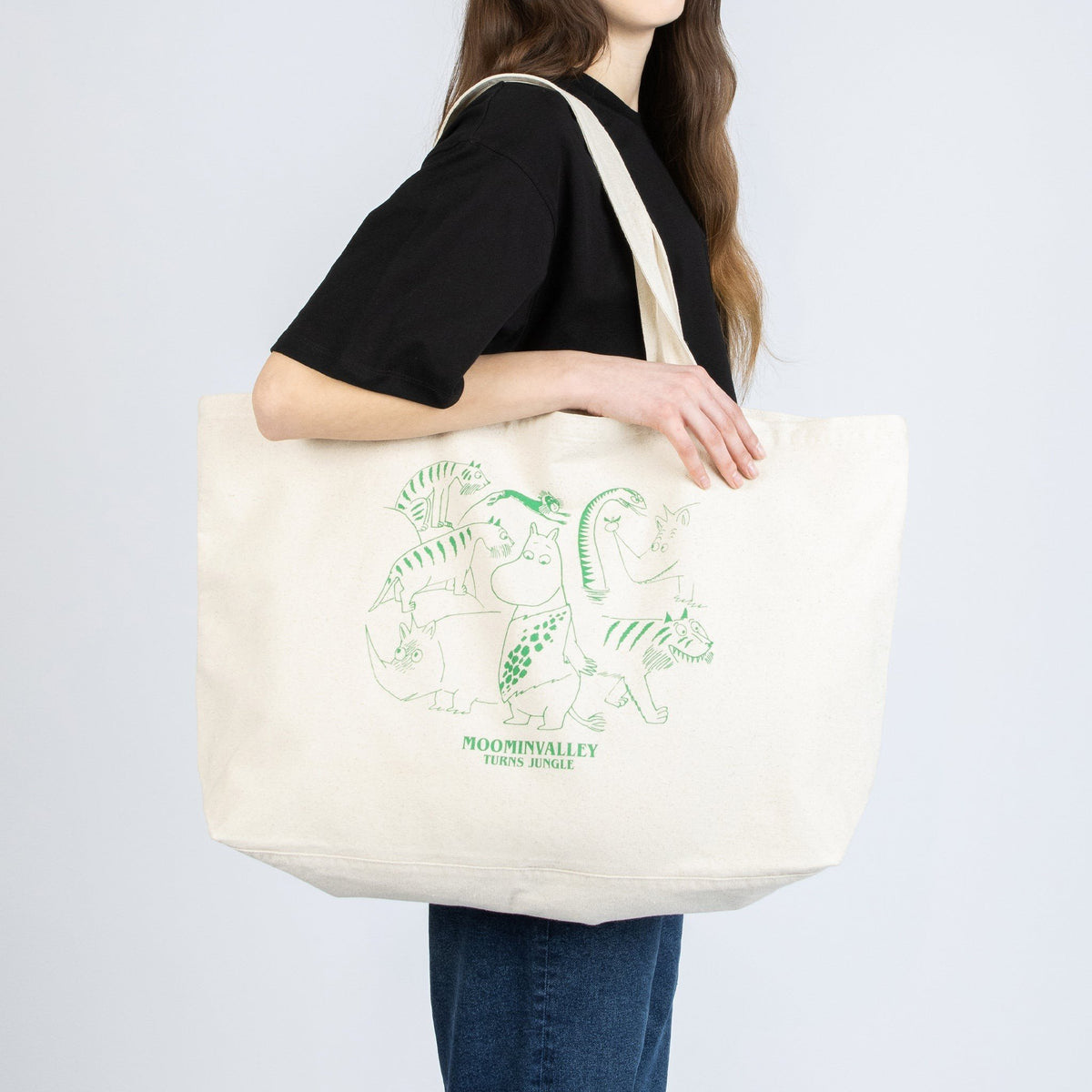1. Woman carrying Moomin Jungle XXL tote bag in off-white, featuring playful Moomin design, studio setting
