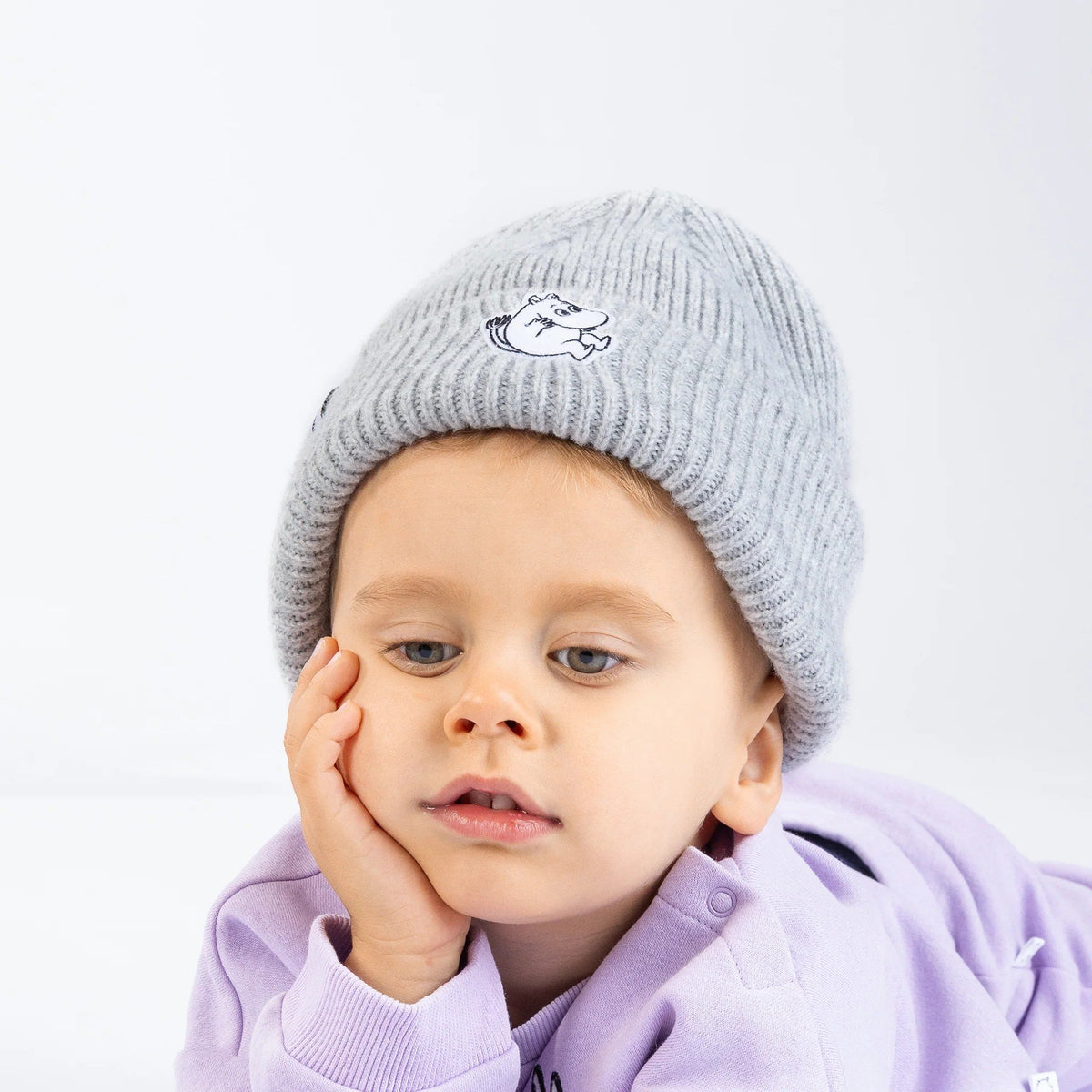 1. Child wearing grey Moomintroll beanie with felt patch, resting head on hand, in a studio setting