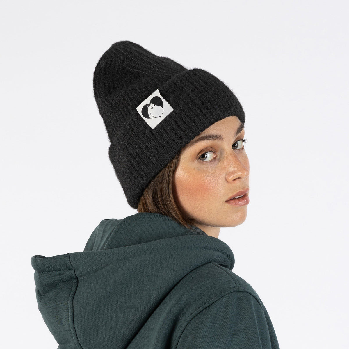 1. Woman wearing black Moomintroll beanie with logo, looking over shoulder, in casual setting