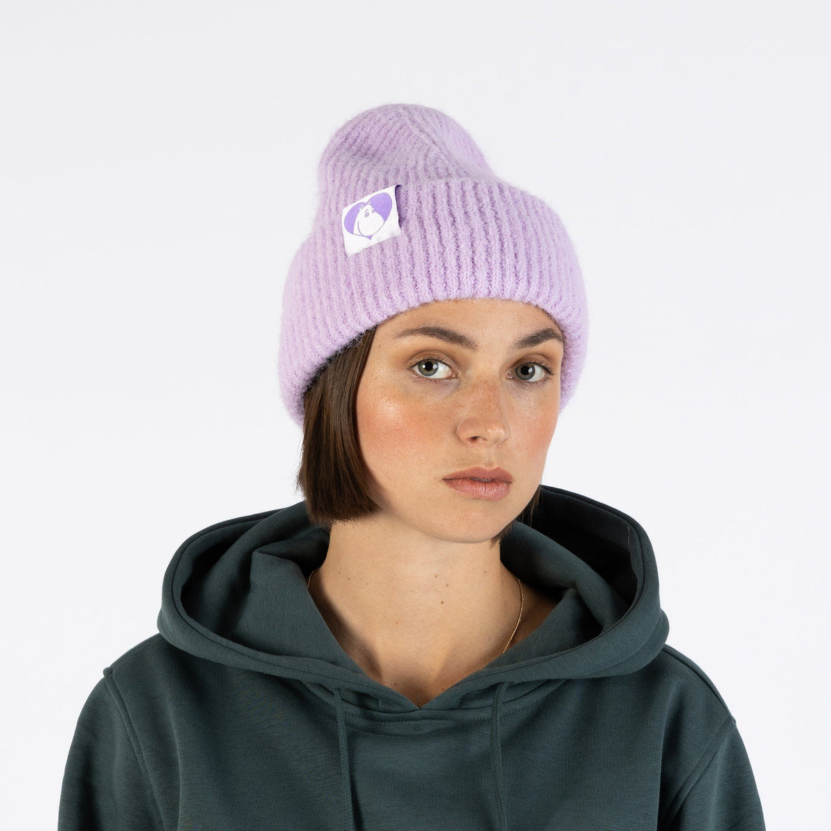 1. Woman wearing lilac Moomintroll beanie with Moomin logo, paired with dark green hoodie, against white background