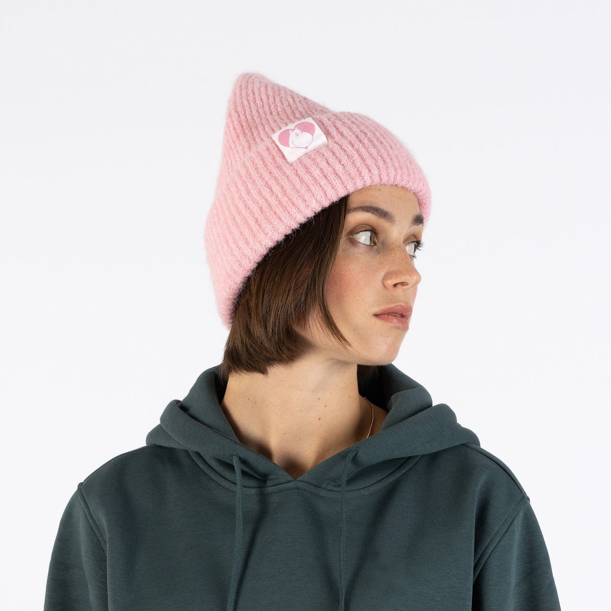 1. Woman in profile wearing pink Moomintroll fluffy chunky beanie by NordicBuddies with logo, paired with green hoodie, studio setting