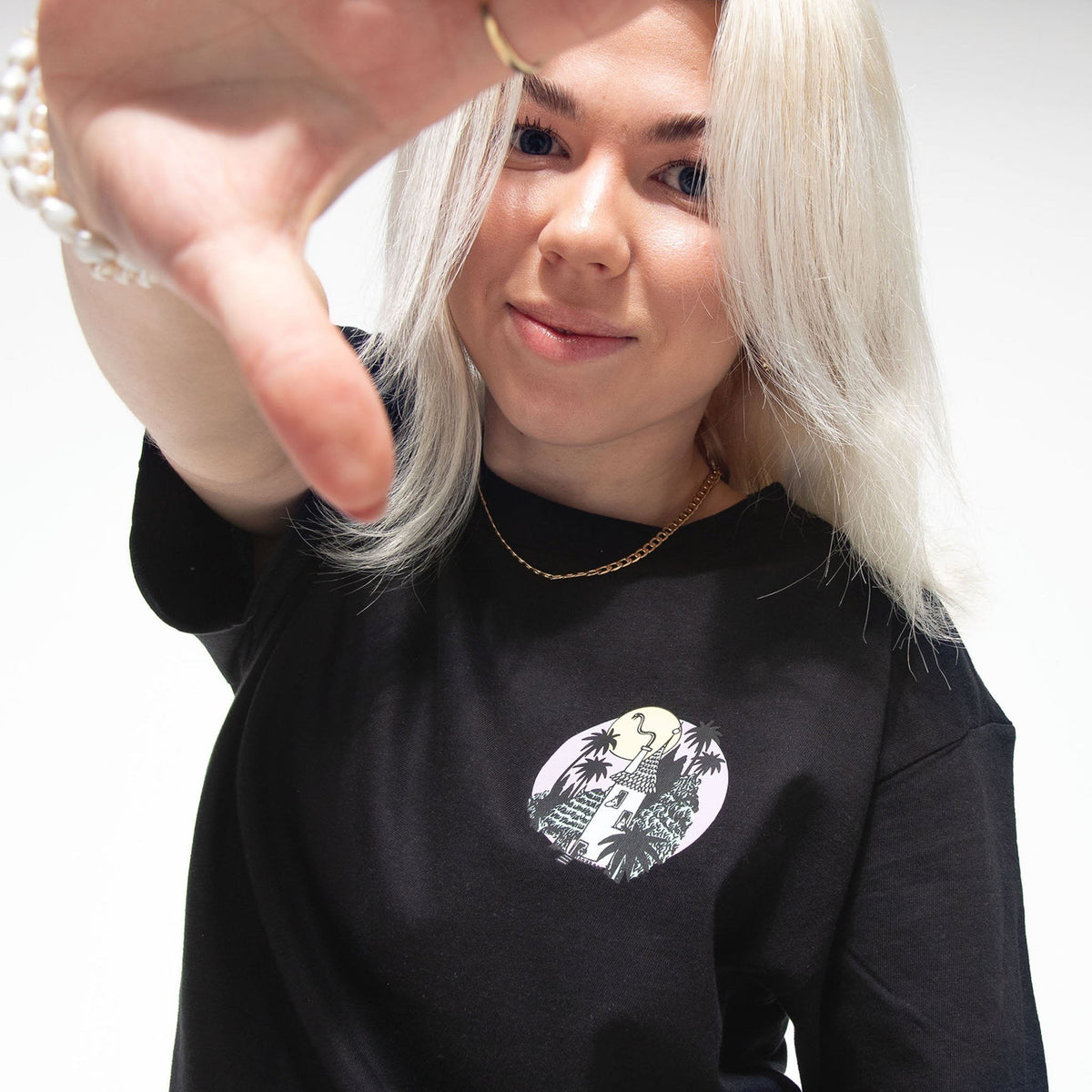 1. Woman wearing black Moomin unisex t-shirt with small front logo, reaching towards camera in studio setting