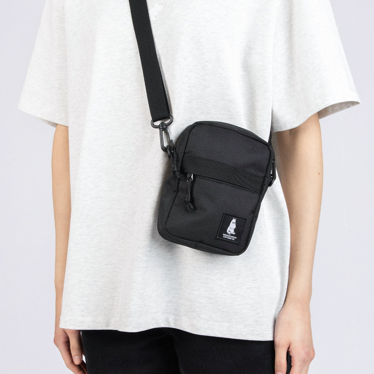1. Person wearing black Moomin by NordicBuddies cross body bag with adjustable strap, featuring Moomin logo, over light grey shirt