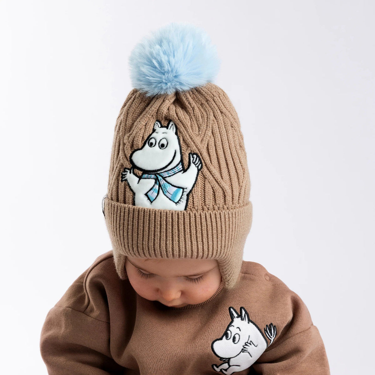 1. Child wearing brown Moomintroll winter beanie with blue pom-pom and fleece lining, featuring Moomintroll character, in studio setting