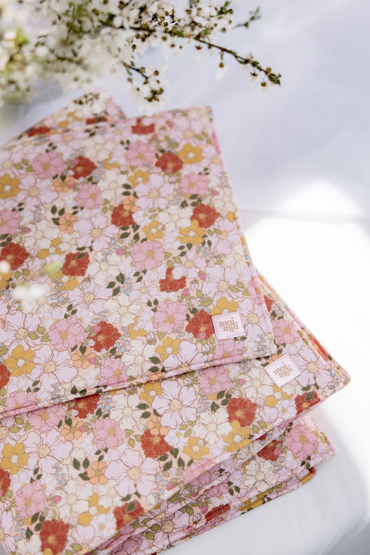 1. Floral muslin baby blanket and pillow set in pink, yellow, and white on a white surface with flowers in the background