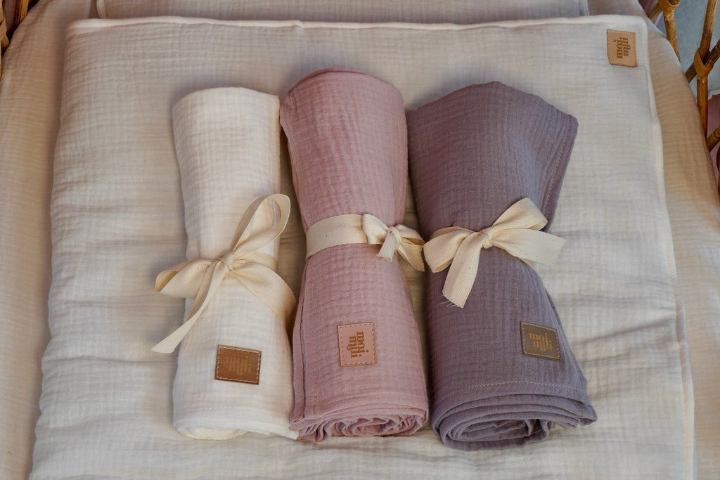 1. Three rolled muslin blankets in cream, pink, and grey tied with ribbons on a cream blanket