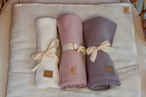 1. Three rolled muslin blankets in cream, pink, and grey tied with ribbons on a cream blanket