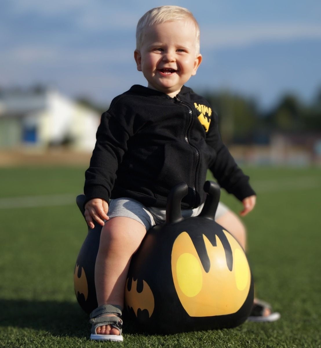 1. Smiling toddler in black hoodie sitting on black and yellow JUMPY Batmobile toy on grass field