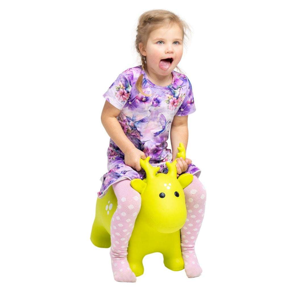 1. Young girl in a floral dress and pink polka dot tights riding a green moose-shaped hopper toy indoors, smiling and having fun
