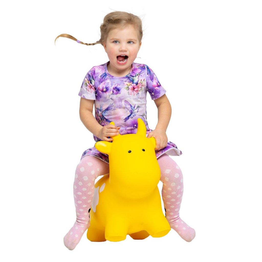 1. Young girl in a purple floral dress and pink polka dot tights sitting on a yellow cow-shaped hopper toy by Gerardo's Toys, smiling with excitement
