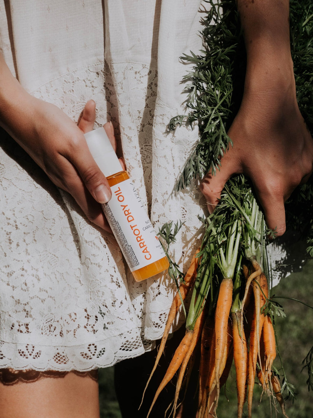 3. Close-up of woman holding LUUV Carrot Dry Oil with fresh carrots, wearing a lace dress