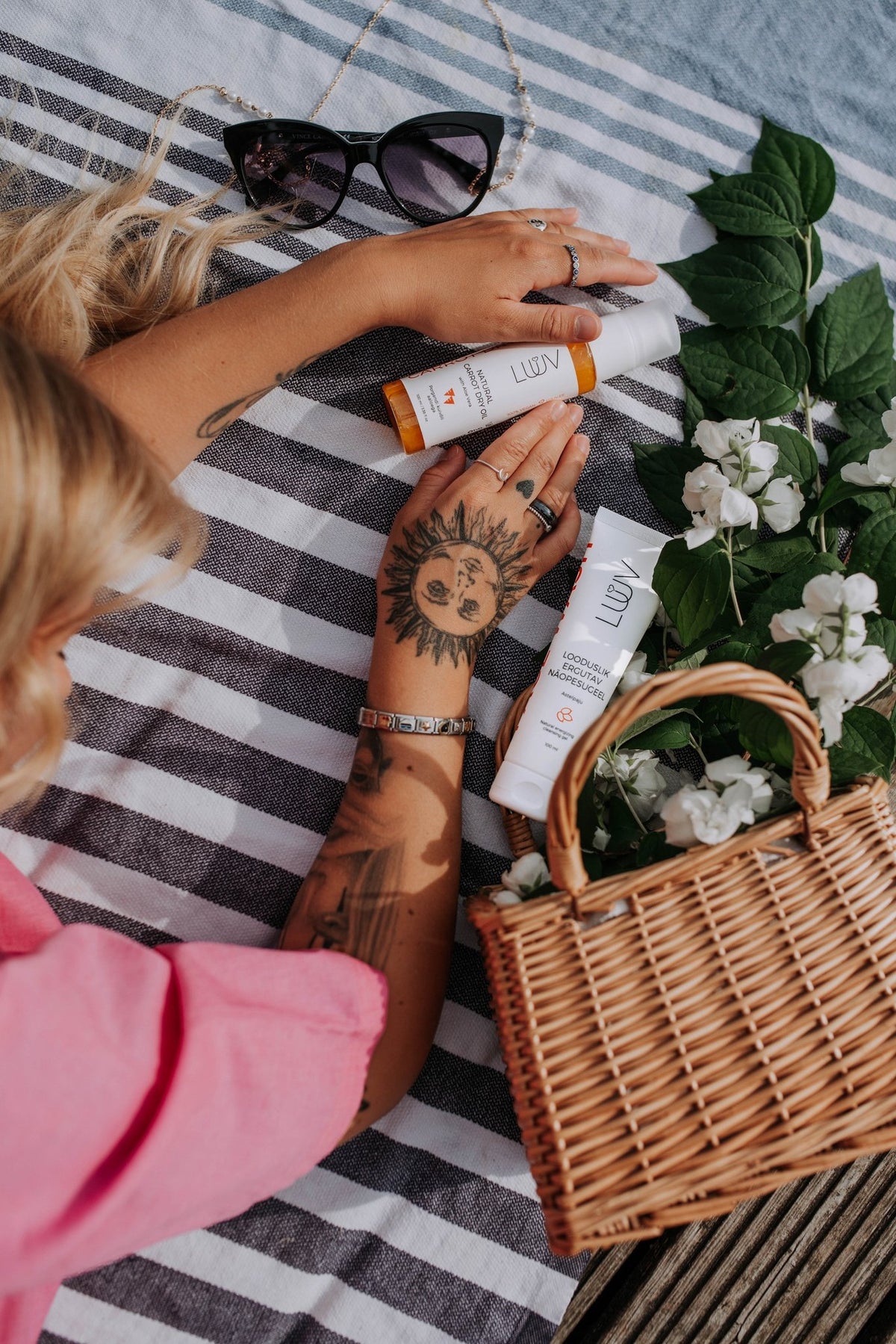 1. Woman lying on striped towel with LUUV Carrot Dry Oil and other skincare products, surrounded by flowers and a wicker basket