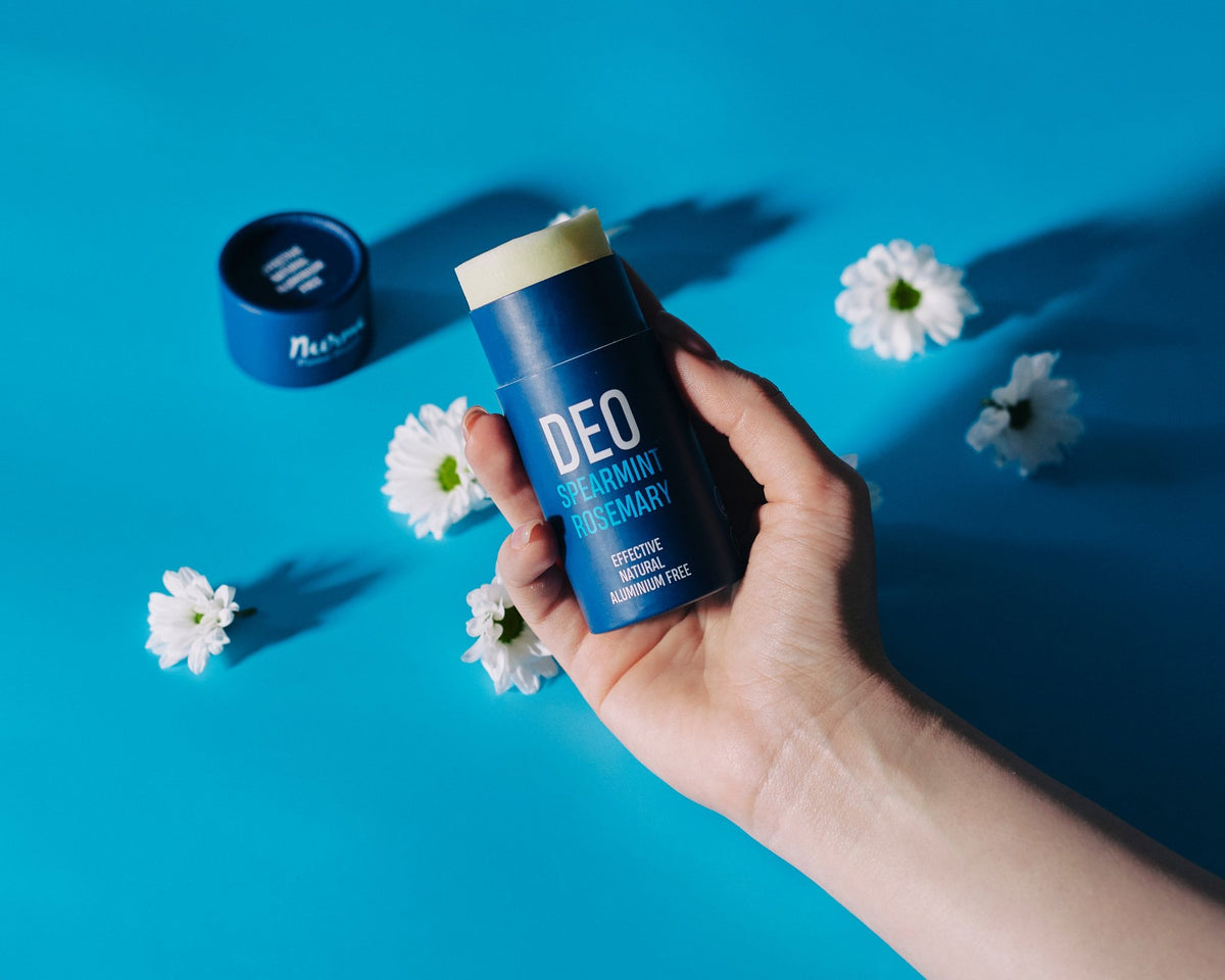 1. Hand holding Nurme spearmint rosemary natural deodorant against blue background with white flowers