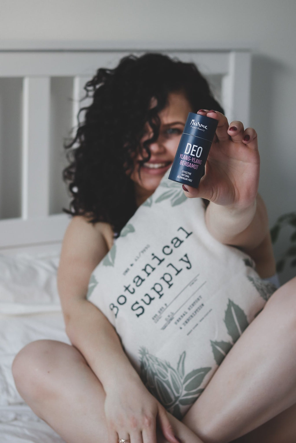 2. Woman holding Nurme Ylang-Ylang Bergamot deodorant while sitting on bed with botanical pillow