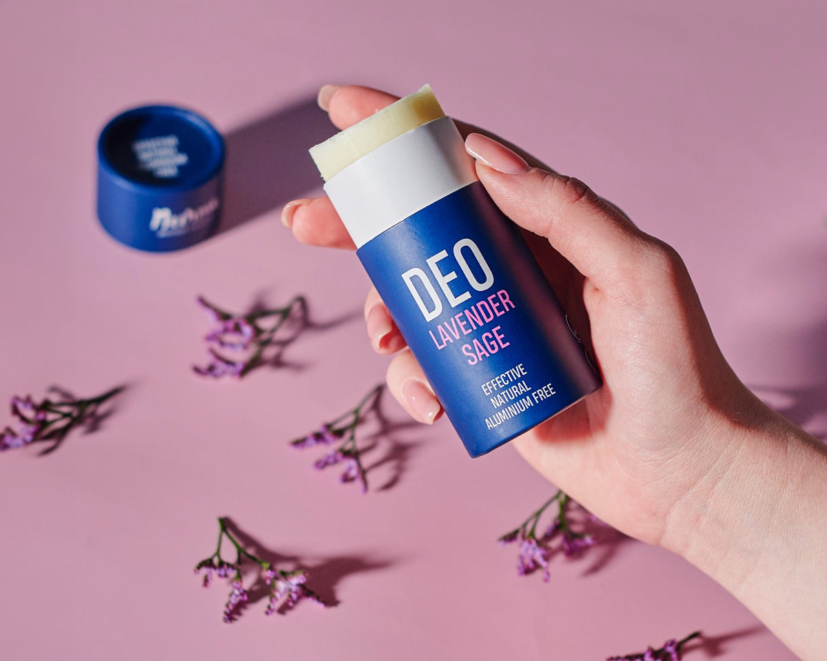 1. Hand holding Nurme Lavender Sage deodorant with open cap and lavender flowers on a pink background