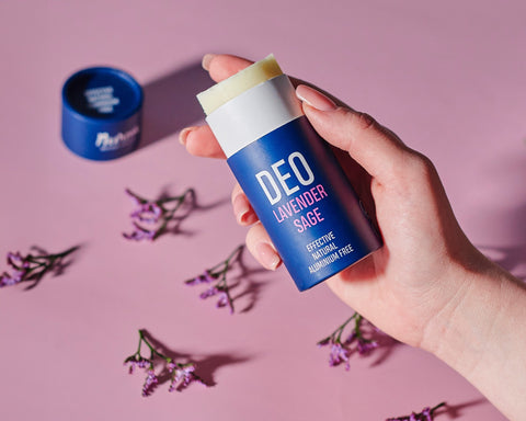 1. Hand holding Nurme Lavender Sage deodorant with open cap and lavender flowers on a pink background