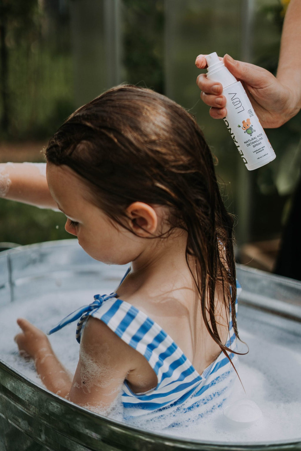 1. Child in blue striped swimsuit in metal tub being sprayed with LUUV Natural Kid's Detangler Spray outdoors