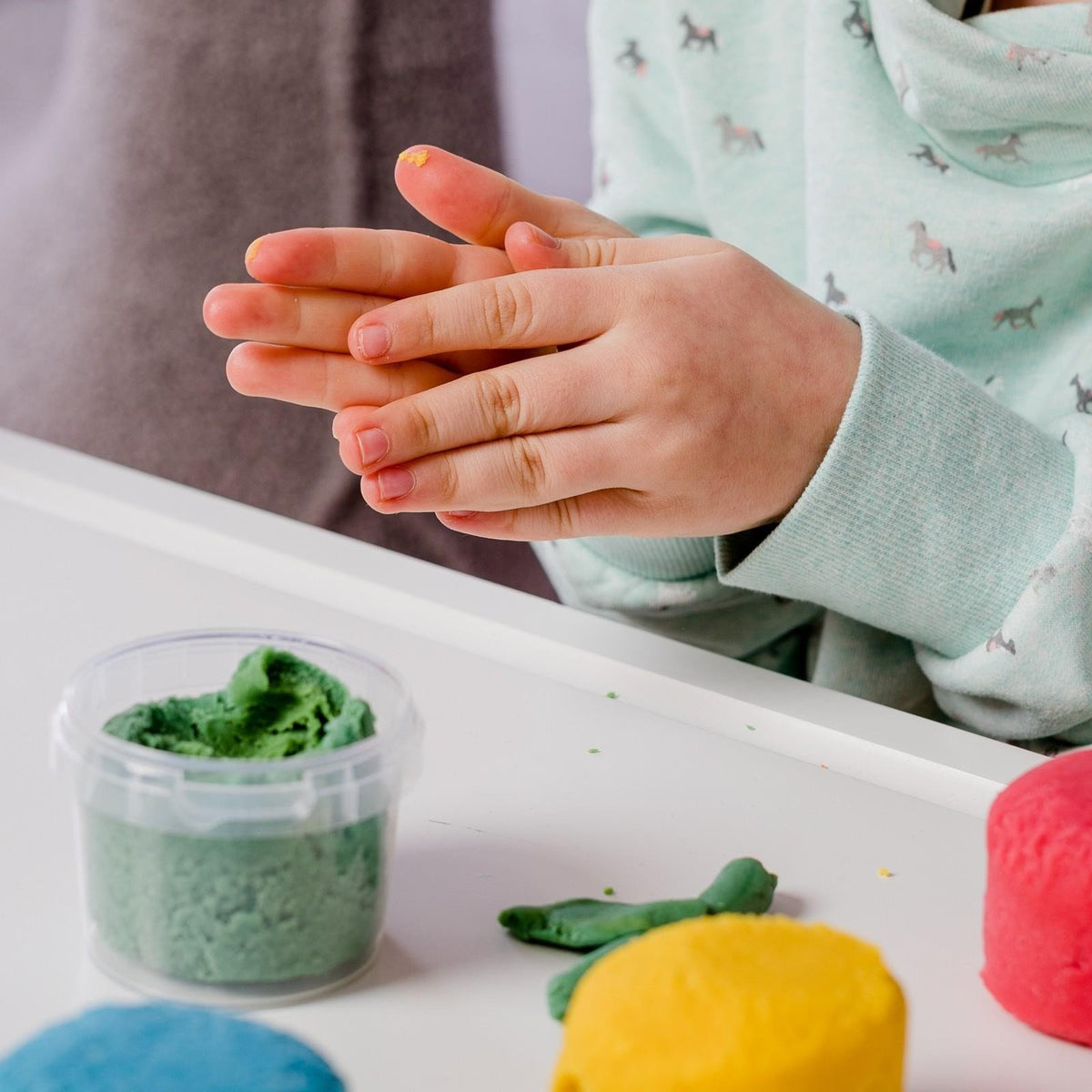 3. Child's hands molding green Neogrün Easy Clay on a white table with other clay colors visible