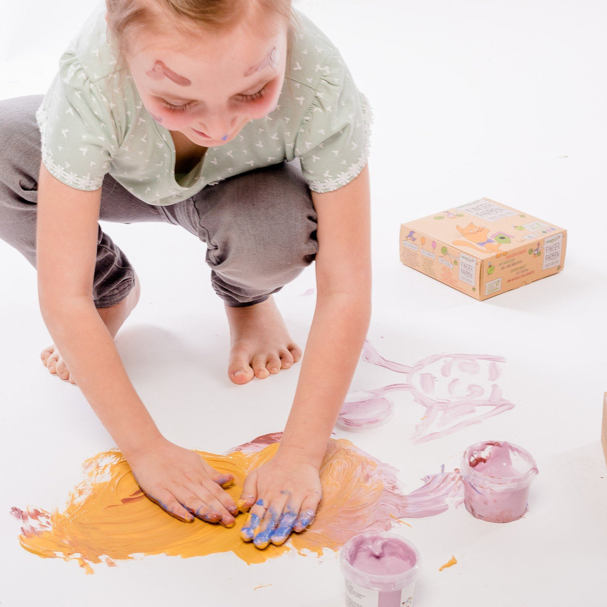 1. Child playing with Neogrün finger paints on white surface, using hands to spread yellow and pink paint, with jars and packaging visible