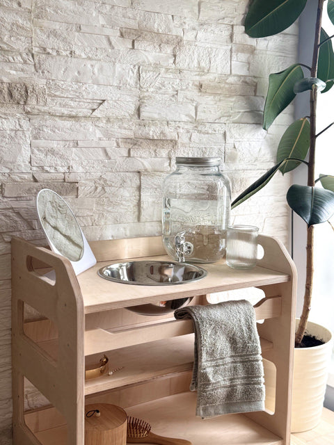 2. Close-up of Montessori washbasin with steel bowl, towel, and glass jar, placed indoors with a stone wall background